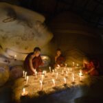 Young Buddhist novices lighting candles by a reclining Buddha statue during Visakha Bucha in Thailand