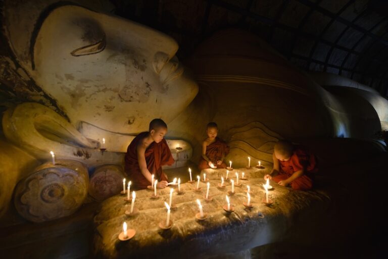 Young Buddhist novices lighting candles by a reclining Buddha statue during Visakha Bucha in Thailand