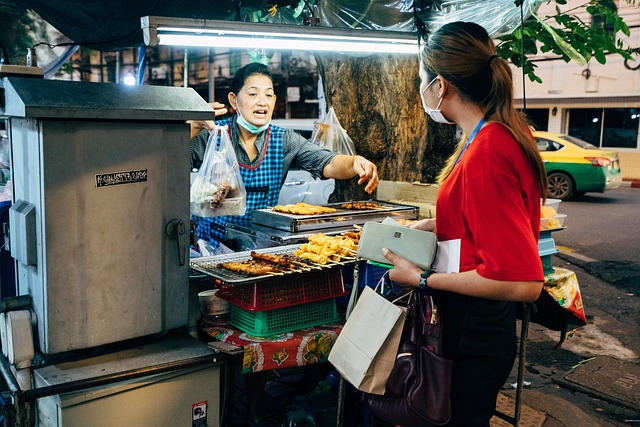 Thai street food vendor handing a bag of snacks to a masked woman at night in Bangkok