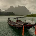 Two longtail boats moored in the rain on a lake in Thailand, with misty mountains in the background during monsoon season