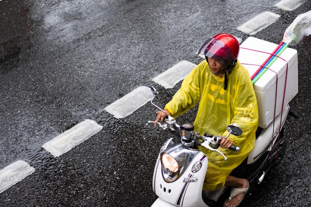 Thai scooter driver wearing a yellow rain poncho, riding on a wet road with a delivery box