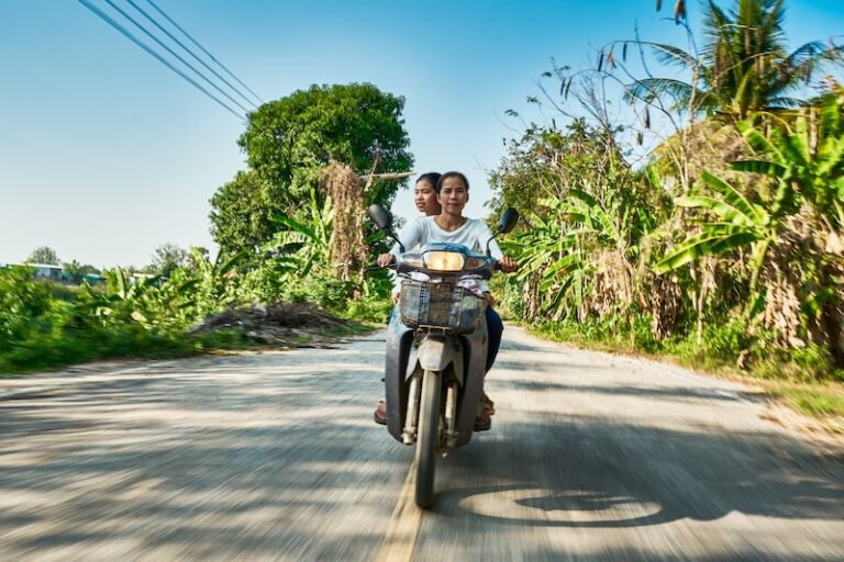 Two Thai women riding a scooter on a rural road surrounded by banana trees and greenery