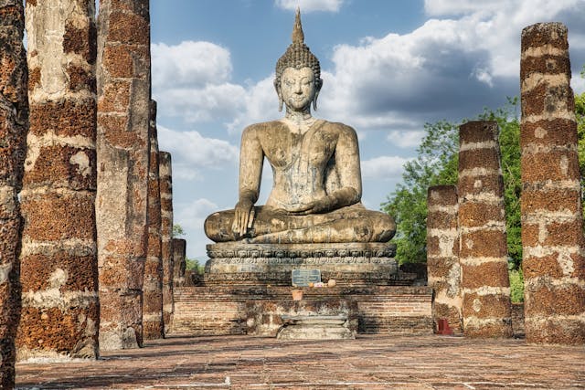 Ancient seated Buddha statue surrounded by stone columns at Sukhothai Historical Park, Thailand, under a partly cloudy sky