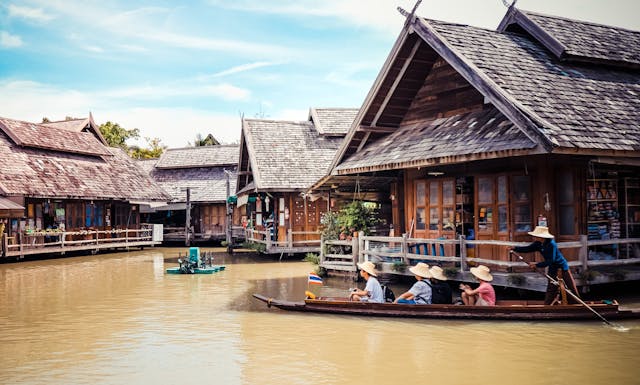 Traditional wooden houses on stilts along a canal in Thailand, with people paddling a boat under a blue sky
