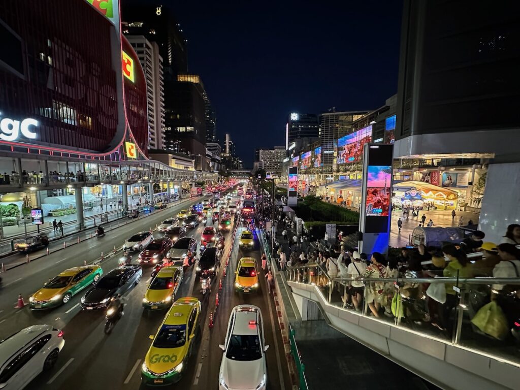 View of a busy Bangkok street at night with colorful neon lights, traffic, taxis, and pedestrians on a skywalk.
