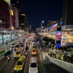 View of a busy Bangkok street at night with colorful neon lights, traffic, taxis, and pedestrians on a skywalk.