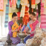 Two smiling Thai women in traditional silk dresses preparing colorful flags and lanterns for a festival, demonstrating a key aspect of Thai culture and tradition.