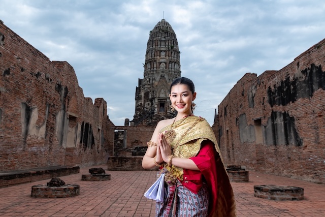 A smiling Thai woman in a traditional costume performs the Wai, the respectful Thai greeting, in front of ancient temple ruins, illustrating a key part of Thai etiquette.