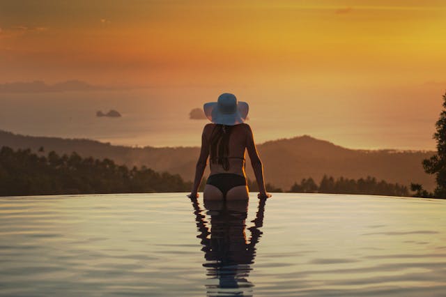 A traveler with a hat relaxes in an infinity pool, watching the sunset over the sea in Thailand, embodying the peace of mind that good travel insurance provides.