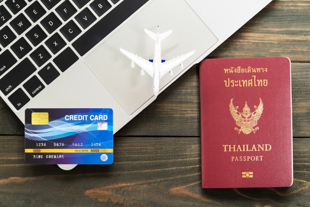 A credit card and a Thai passport next to a laptop and a model airplane, illustrating the process of booking a trip and the need for credit card travel protection for Thailand.