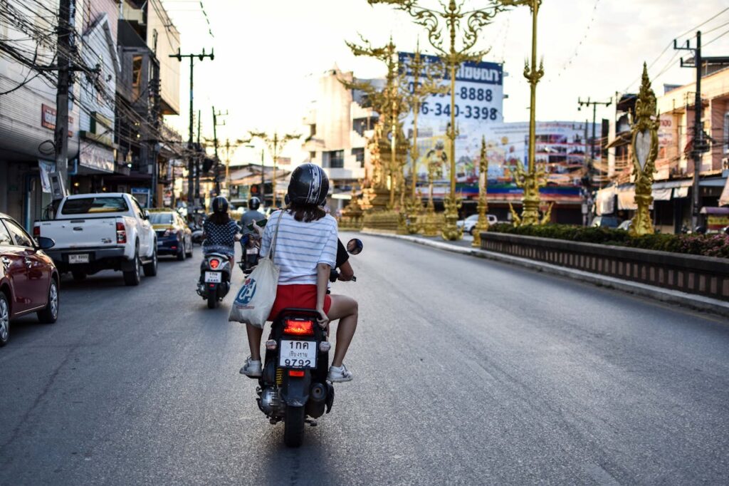 Two travelers riding a scooter in Thailand traffic, highlighting the risk of road accidents for tourists.