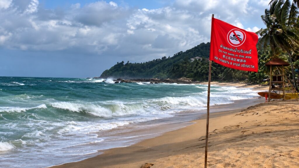 Red warning flag prohibiting swimming on a beach in Thailand, with strong waves in the background.