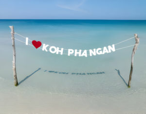 Banderole 'I ♥ KOH PHANGAN' entre deux poteaux sur une plage tranquille de Koh Phangan avec l'ombre reflétant sur l'eau cristalline.