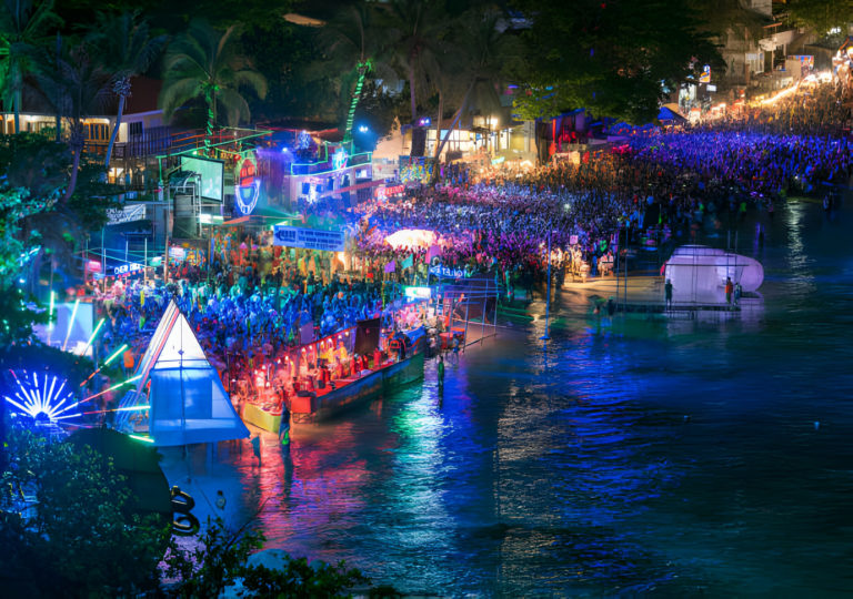 Foule colorée lors de la Full Moon Party de nuit sur la plage de Haad Rin à Koh Phangan, Thaïlande, avec des jeux de lumière et reflets sur l'eau.