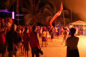 Fête nocturne sur une plage avec des participants dansant sous des lumières néon et une décoration tropicale à Koh Phangan, Thaïlande.