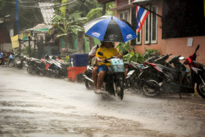 Livreur à moto sous la pluie tenant un parapluie ouvert, naviguant dans une rue inondée.