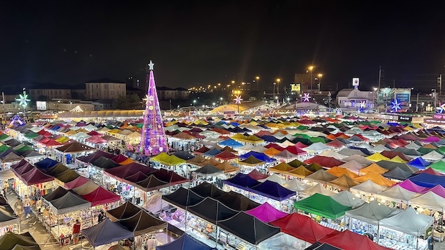 Vue aérienne d'un grand marché de nuit à Bangkok, montrant des centaines de stands aux toits de tentes multicolores et un grand arbre de Noël illuminé.