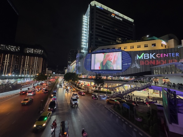 Vue nocturne du centre commercial MBK Center à Bangkok, avec son grand écran illuminé et le trafic routier sur l'avenue.
