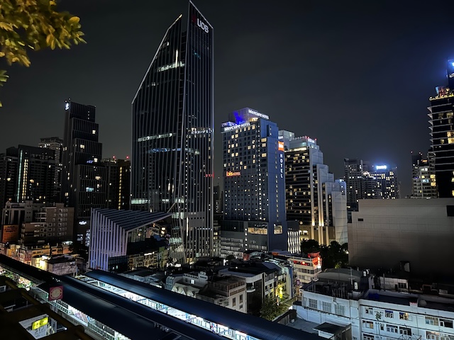Vue nocturne sur les gratte-ciels illuminés de Bangkok, prise depuis le toit du centre commercial EmQuartier, avec la tour UOB et la station de BTS au premier plan.