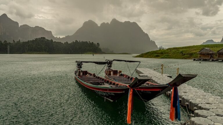 Deux bateaux traditionnels amarrés sur un lac paisible en Thaïlande sous une pluie fine, entourés de montagnes et d’un ciel brumeux