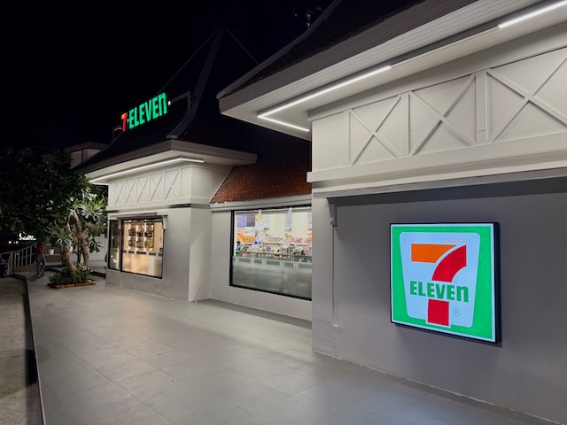 Vue latérale d'un bâtiment 7-Eleven en Thaïlande au design moderne blanc, avec une grande enseigne carrée et lumineuse sur le mur et l'intérieur du magasin visible par la fenêtre.