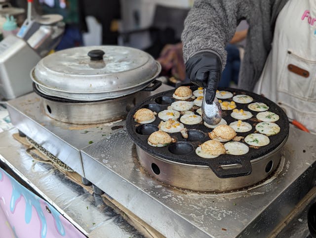Préparation du Kanom Krok, un snack thaïlandais sucré, dans un moule en fonte sur un stand de street food.