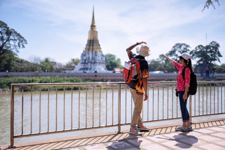 Deux touristes en sac à dos face à un temple thaïlandais au bord d’une rivière