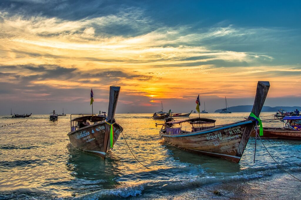 Deux bateaux long-tail traditionnels thaïlandais au premier plan, flottant sur l'eau au coucher du soleil sur la plage d'Ao Nang à Krabi, avec un ciel spectaculaire orange et bleu.