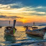 Deux bateaux long-tail traditionnels thaïlandais au premier plan, flottant sur l'eau au coucher du soleil sur la plage d'Ao Nang à Krabi, avec un ciel spectaculaire orange et bleu.
