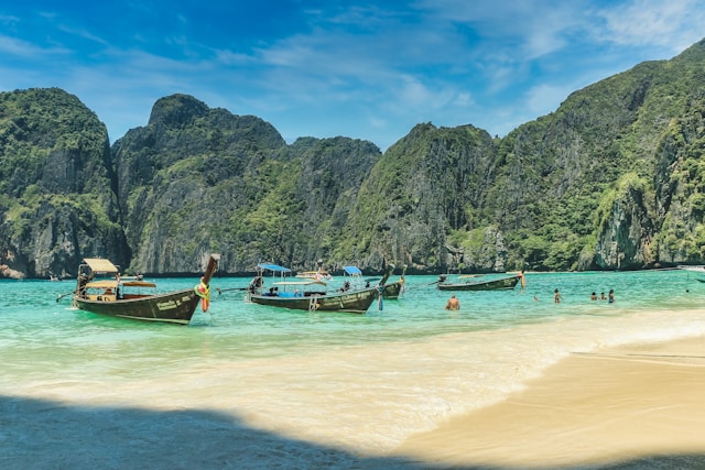 Vue de la plage de Maya Bay à Koh Phi Phi avec des bateaux long-tail dans une eau turquoise et des falaises calcaires en arrière-plan.