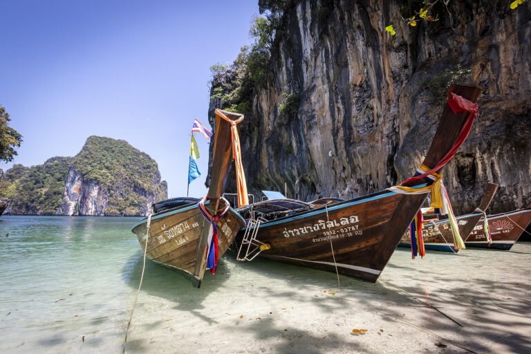 Deux bateaux à longue queue en bois avec des rubans colorés, amarrés sur le sable d'une plage thaïlandaise, face à une mer turquoise et une falaise calcaire.