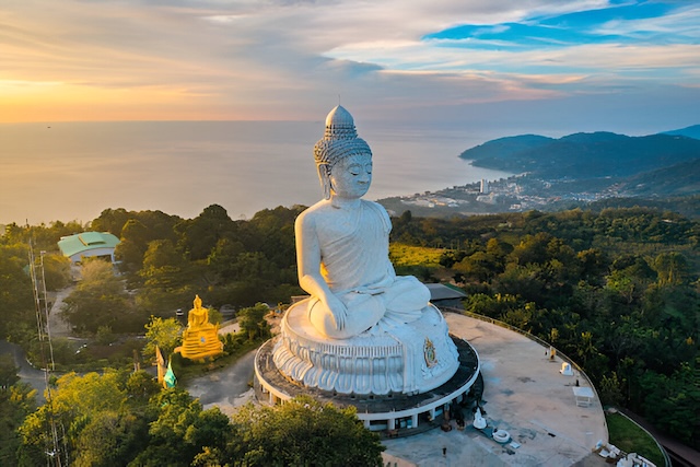 Vue aérienne de la statue du Grand Bouddha de Phuket en marbre blanc, dominant les collines verdoyantes et la baie de Karon au coucher du soleil.