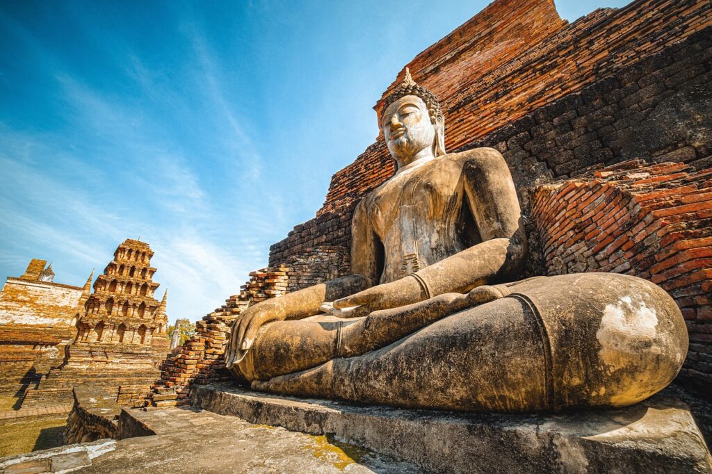 Vue en contre-plongée d'une grande statue de Bouddha en pierre, assise devant les ruines en briques du temple Wat Mahathat à Sukhothai, sous un ciel bleu.
