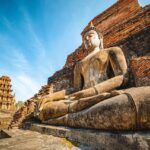 Vue en contre-plongée d'une grande statue de Bouddha en pierre, assise devant les ruines en briques du temple Wat Mahathat à Sukhothai, sous un ciel bleu.