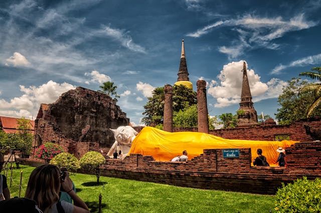 Statue du grand Bouddha couché recouvert de sa robe safran au temple Wat Yai Chai Mongkhon, avec les ruines et le chedi en arrière-plan.