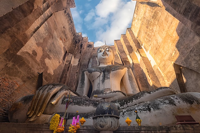 Vue en contre-plongée du Bouddha géant en pierre du Wat Si Chum, assis à l'intérieur de son mondop (bâtiment sans toit), avec un ciel bleu visible au-dessus.