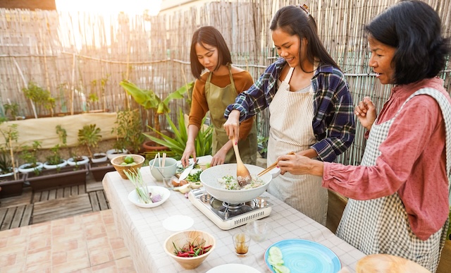 Trois femmes asiatiques souriantes cuisinant ensemble en plein air sur une terrasse, préparant un plat dans un wok blanc sur un réchaud.