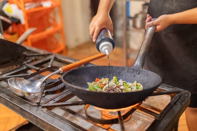 Main d'un chef versant de la sauce sur des légumes frais qui cuisent dans un wok sur un feu vif lors d'un cours de cuisine en Thaïlande.
