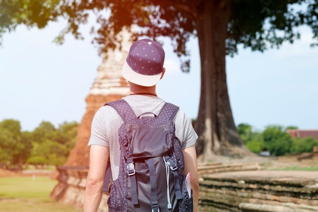 Voyageur de dos avec un sac et une casquette, marchant vers un ancien temple en brique dans le parc historique d'Ayutthaya en Thaïlande.