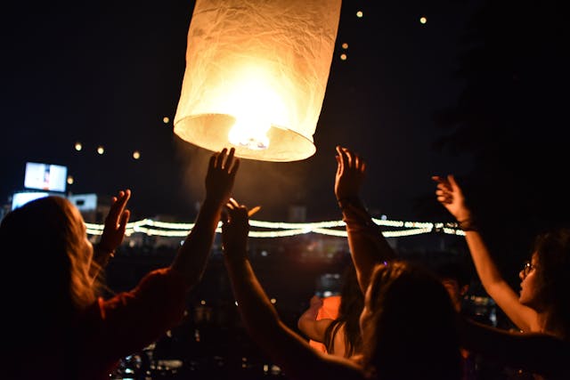 Des mains de personnes lèvent une lanterne céleste illuminée dans le ciel sombre lors du festival de Loy Krathong en Thaïlande, avec d'autres lanternes en arrière-plan.