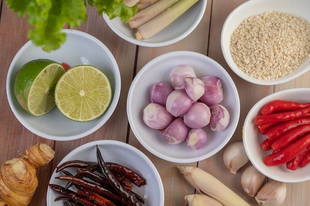 Assortiment d'ingrédients frais pour la cuisine thaïlandaise, incluant piments, citronnelle, galanga, échalotes et citron vert, disposés dans des bols sur une table en bois.