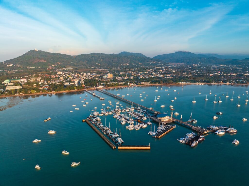 Vue aérienne de la marina de Chalong à Phuket, remplie de yachts et de bateaux d'excursion, avec la jetée s'avançant dans la mer et le Grand Bouddha visible sur les collines.