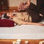 Cliente souriante recevant un étirement de la jambe par un praticien lors d'une séance de massage thaïlandais traditionnel sur un matelas au sol.