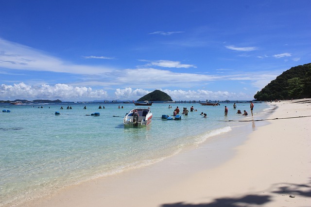 Baigneurs et bateau rapide sur une plage de sable blanc à Phuket, avec une île verdoyante en arrière-plan sous un ciel bleu.