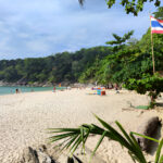 Plage de sable blanc en Thaïlande avec drapeau thaïlandais, mer turquoise et vacanciers, entourée de végétation tropicale