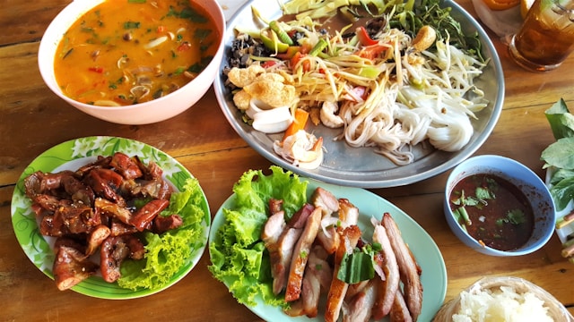 Table en bois avec un repas complet de cuisine Isan thaïlandaise, incluant une salade de papaye Som Tum, du porc grillé Kor Moo Yang, une soupe Tom Yum et du riz gluant.