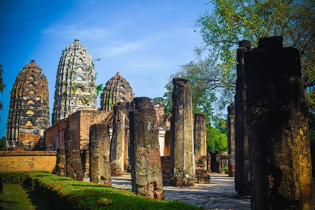 Vue des ruines du temple Wat Mahathat à Sukhothai, avec des piliers en pierre au premier plan et des tours-reliquaires (prangs) en brique et en stuc à l'arrière-plan, sous un ciel bleu.
