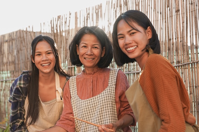 ortrait de trois femmes thaïlandaises de générations différentes (une mère et ses deux filles) souriant chaleureusement à la caméra, portant des tabliers devant une clôture en bambou.