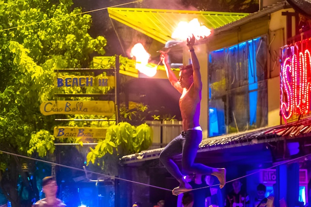 Jongleur de feu torse nu réalisant une performance acrobatique la nuit à Koh Phi Phi, avec des enseignes de bar illuminées en arrière-plan.