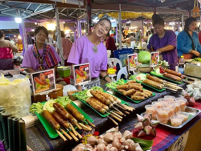 Vendeuses thaïlandaises souriantes sur un stand de nourriture vendant des brochettes de saucisses grillées et des spécialités locales sur un marché de nuit à Ayutthaya.
